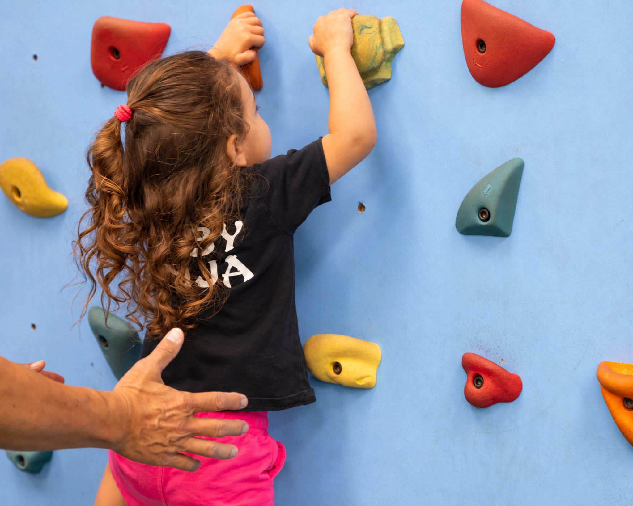 Young child climbing an indoor rock wall with help from a caregiver during a Lil Ninjas NinjaZone class at Impact Sports Center in Lodi.